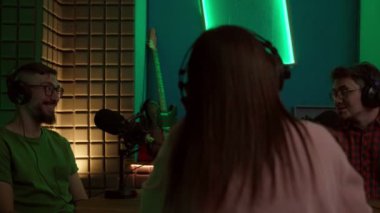 Modern stüdyoda podcast yaparken mikrofonlu masada oturan ve iletişim kuran gündelik kıyafet ve kulaklıklar takan konsantre genç erkek ve bayan meslektaşlar.