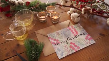 Renkli kartpostallı Noel kompozisyonunun en üst görüntüsü üzerinde Feliz Navidad yanan mumların yanına ve renkli bitki dallarıyla süslenmiş ahşap masa üzerindeki çay fincanlarına yerleştirilmiş.