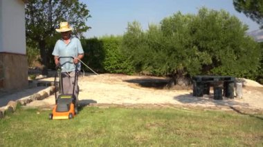 Full body of male gardener in hat mowing grassy lawn near bushes and trees in summer