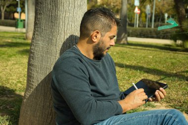 Güneşli bir günde açık havada dijital tablet kullanan mavi tişörtlü, gülen sakallı bir şehirli. Bağlantı ve kablosuz teknoloji