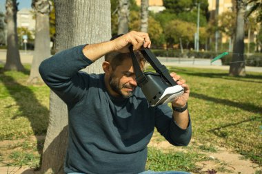 Gülümseyen mavi gömlekli genç adam, VR kulaklığını ayarlıyor, doğal arka planda duruyor.