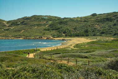 MENORCA, Balearik Adaları, 16 Nisan 2022: Güneşli bir günde Cala Binimela, Cami de Cavalls rotası çizen birkaç bisikletçi. Bir sürü bitki örtüsü olan deniz ve dağ manzarası