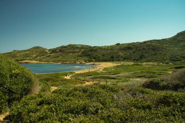 MENORCA, Balearik Adaları, 16 Nisan 2022: Güneşli bir günde Cala Binimela, Cami de Cavalls rotası çizen birkaç bisikletçi. Bir sürü bitki örtüsü olan deniz ve dağ manzarası