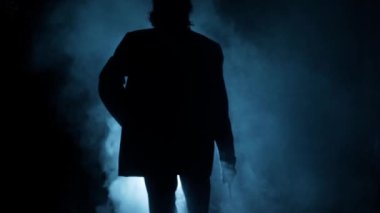 Elinde silahla dolaşan takım elbiseli bir gangsterin dramatik sinematik çekimi. Dramatik ışıkta yürüyen bir gangsterin siluetleri, duman bulutları. Yüksek çözünürlüklü görüntü. Yavaş Hareket.