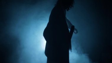 Mavi takım elbiseli bir adamın elinde balta ve iple dramatik bir ışığın önünden geçerken görüntüsü..