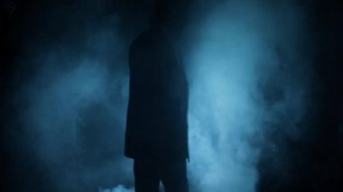 Mavi takım elbiseli bir adamın elinde balta ve iple dramatik bir ışığın önünden geçerken görüntüsü..