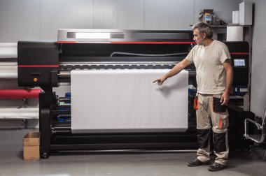 Teknisyen, dijital yazıcı ofisindeki büyük endüstriyel yazıcı ve çizer makinesinde beyaz kağıdın boş alanını gösteriyor ve gösteriyor.