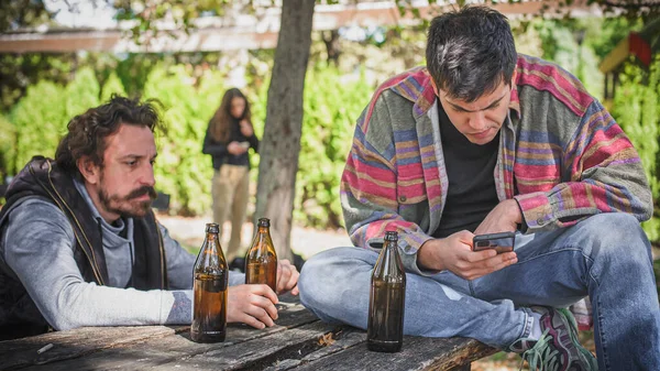Fiatalember Iszik Sört Palackból Barátaival Nézd Meg Mobiltelefonját Üljön Padon — Stock Fotó