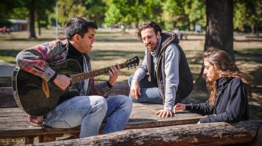 Pozitif ve rahat genç arkadaşlar parkta bankta otururken birlikte gitar çalıp şarkı söylüyorlar ve güzel romantik yaz gününün tadını çıkarıyorlar.
