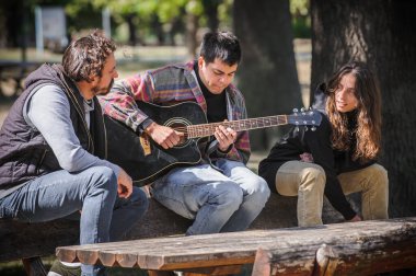 Pozitif ve rahat genç arkadaşlar parkta bankta otururken birlikte gitar çalıp şarkı söylüyorlar ve güzel romantik yaz gününün tadını çıkarıyorlar.