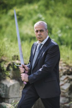 Portrait of handsome businessman model man in a modern casual suit with a large medieval sword. Modern fashion street style concept. Elegant urban warrior. Corporate and money security