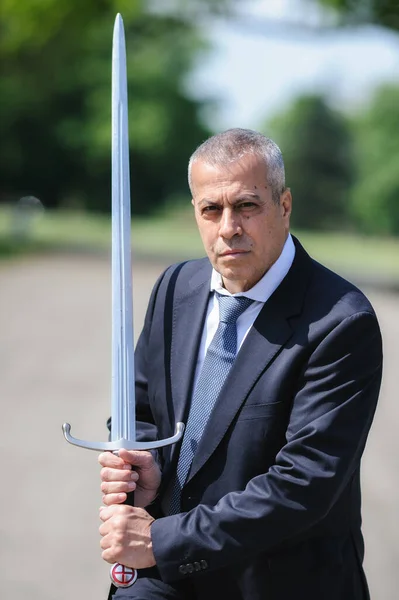 Portrait of handsome businessman model man in a modern casual suit with a large medieval sword. Modern fashion street style concept. Elegant urban warrior. Corporate and money security