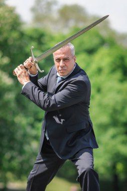 Portrait of handsome businessman model man in a modern casual suit with a large medieval sword. Modern fashion street style concept. Elegant urban warrior. Corporate and money security