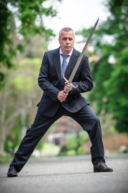 Portrait of handsome businessman model man in a modern casual suit with a large medieval sword. Modern fashion street style concept. Elegant urban warrior. Corporate and money security