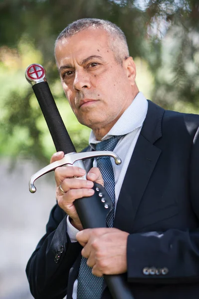 Portrait of handsome businessman model man in a modern casual suit with a large medieval sword. Modern fashion street style concept. Elegant urban warrior. Corporate and money security