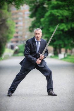 Portrait of handsome businessman model man in a modern casual suit with a large medieval sword. Modern fashion street style concept. Elegant urban warrior. Corporate and money security