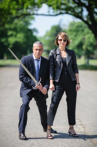Portrait of handsome businessman model man in a modern casual suit protect woman love with a large medieval sword. Modern fashion street style concept. Elegant urban warrior couple