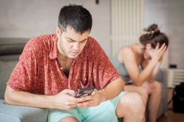 Genç üzgün bir kadın, biriyle telefonda mesajlaşan ve onunla ilgilenmeyen ortağının yanında..