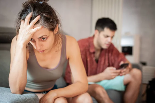 A young sad woman is next to her partner who is texting on the phone with someone, and not paying attention to her.