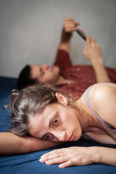 A young sad woman is next to her partner who is texting on the phone with someone, and not paying attention to her.