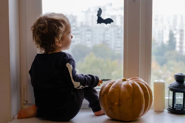 Een kleine jongen in skelet kostuum en heksenhoed voor Halloween zit voor het raam en lacht — Stockfoto