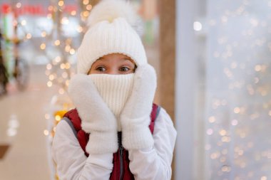 Alışveriş merkezinde beyaz şapkalı ve fularlı bir çocuk şaşırdı. Arkaplanda bulanık parlak ışıklar