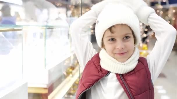 サンタスの帽子の男の子は市場の窓を通って店を調べる。背景に輝く光が. — ストック動画