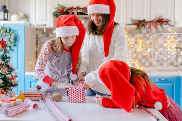 Noel Baba şapkalı kadın ve kızları Noel hediyelerini paketler, makasla kağıt keserler.