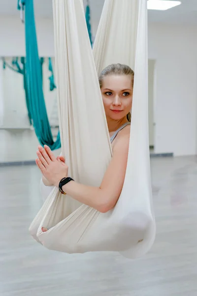 Женщина 20-30 лет висит на стропах белого гамака для воздушной йоги и делает намасте — стоковое фото