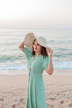 Denize yolculuk. Elbiseli ve şapkalı bir kız kumsalda yürüyor. Deniz kıyısında yürüyen bir turist. Tatil döneminde sıcak seyahatler. Boşluğu kopyala