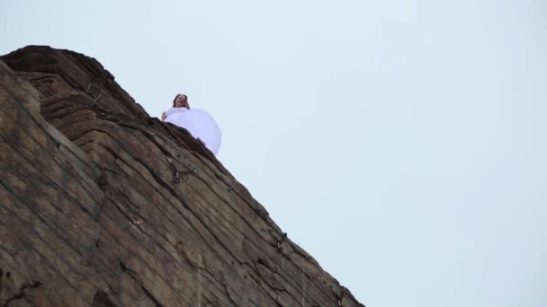 Vue du bas : la mariée sur le bord de la falaise. Séance photo extrême : une fille en robe blanche en hiver — Video