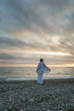 Denize yolculuk. Elbiseli ve şapkalı bir kız kumsalda yürüyor. Deniz kıyısında yürüyen bir turist. Tatil döneminde sıcak seyahatler. Boşluğu kopyala