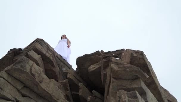 Вид знизу: наречена на краю скелі. екстремальна фотосесія: дівчина в білій сукні взимку — стокове відео
