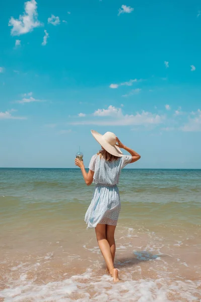 Cesta k moři. dívka v šatech a klobouku kráčí po pláži. turistické procházky podél mořského pobřeží. horké výlety během prázdnin. kopírovat prostor — Stock fotografie