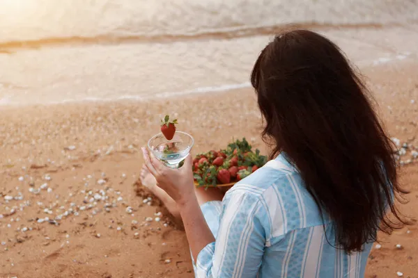 Föda. Hälsosam mat och vila. kvinna dricker rent vatten i glas bakgrund hav och äter jordgubbar. En kvinna på stranden njuter av livet — Stockfoto