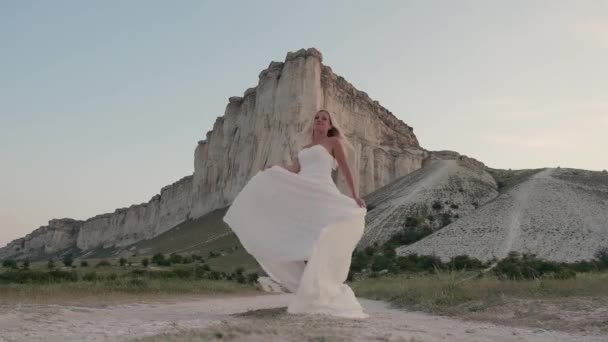Jonge mooie blonde vrouw in een witte jurk draaiend in de berg. Aantrekkelijk modemodel met vliegend haar. Een mooie vrouw schudt haar. Langzame beweging — Stockvideo