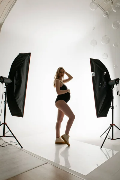 Uma mulher grávida em um terno preto acaricia sua barriga em um fundo branco. Uma rapariga no estúdio sob os holofotes. iluminação profissional. — Fotografia de Stock