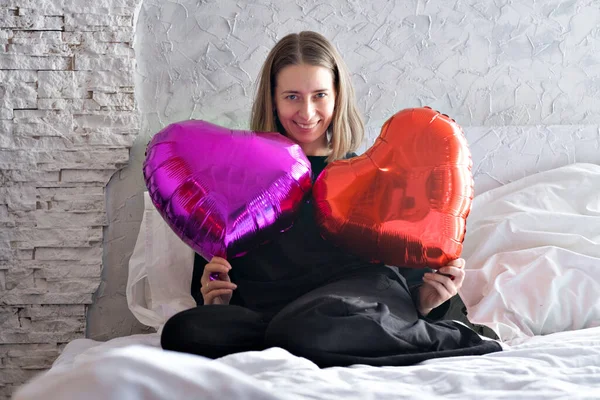 Una mujer en un suéter negro sentada en la cama en el dormitorio con globo rojo en forma de corazón. Amor y San Valentín concepto del día. Imágenes De Stock Sin Royalties Gratis