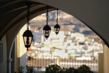 Bir kilisenin çatısında asılı fenerler ve arka planda Fira Santorini köyü.