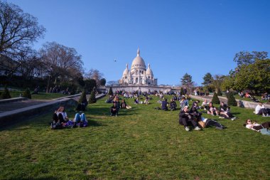 Paris, Fransa - 22 Mart 2022: The Basilica of the Sacred Heart of Paris, namı diğer Sacre - Coeur ve çimlerin üzerinde oturan bir grup insan