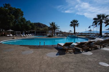 Ios, Yunanistan - 12 Haziran 2021: Yunanistan 'daki Mylopotas plajı önünde güneşli yataklı ve palmiye ağaçlı bir açık havuz manzarası