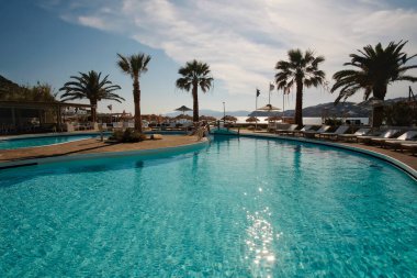 Ios, Yunanistan - 12 Haziran 2021: Yunanistan 'daki Mylopotas plajı önünde güneşli yataklı ve palmiye ağaçlı bir açık havuz manzarası