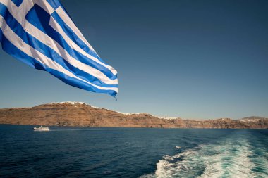 Yunanistan 'daki Santorini adasının önünde rüzgarda dalgalanan dev Yunan bayrağı ve uzakta bir feribot. 