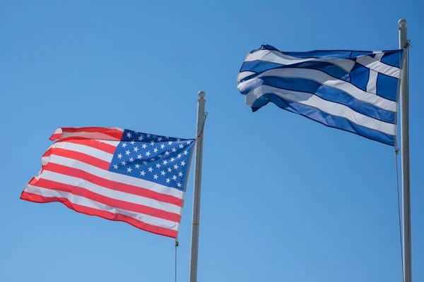 The national flags of the United States of America and Greece waving in the wind and a blue sky in the background