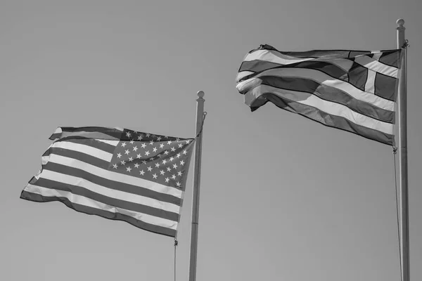 The national flags of the United States of America and Greece waving in the wind and a blue sky in the background in black and white