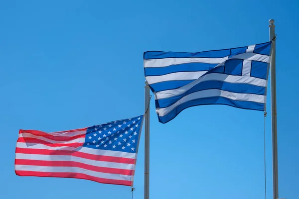 The national flags of the United States of America and Greece waving in the wind and a blue sky in the background