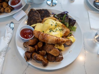 Kahvaltıda bol bol Benedict yumurtası, patates kızartması ve hindi sosisi serpilmiş mermer bir masada.