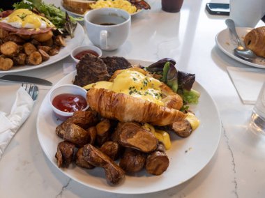 Kahvaltıda bol bol Benedict yumurtası, patates kızartması ve hindi sosisi serpilmiş mermer bir masada.