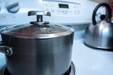 Kapağı açık büyük gümüş bir tencerenin yakın görüntüsü, fırının tepesinde pişmiş bir yemeği sıcak tutarken içinde buhar oluşuyor.