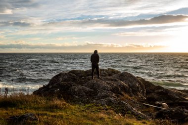 Gün batımında San Juan Adası 'nın kayalık kıyı şeridinde duran siluetli bir adam görülüyor. Arka planda ince beyaz bulutlar var.